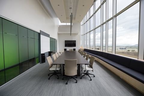 A clean and bright conference room in an office with a flat screen TV, long conference table, 16 chairs around it and big windows that allow sunlight in.