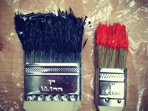 Two paint brushes stained with different color paint. The two inch brush on the left is stained with black paint, and the one inch brush is stained with red paint. 