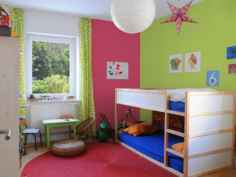 A small kid’s room with a bunk bed, green and pink walls, a white window, green and white curtains and lots of toys.