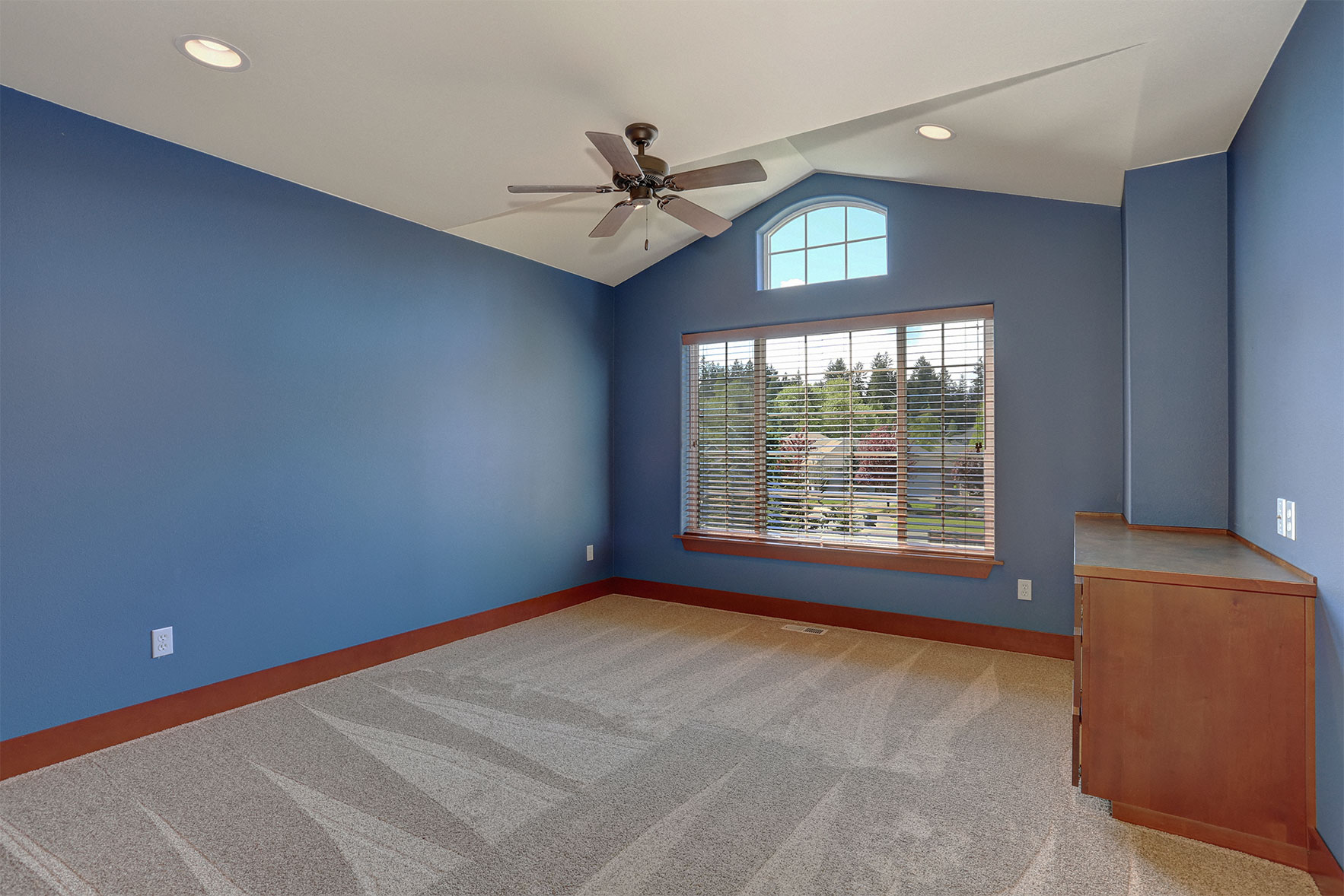 A room that has a ceiling fan, large windows, a grey carpet, wooden cabinet and a fresh coat of blue interior paint.