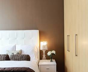 White bed, nightstand with a lamp and flowers on it, and a closet in front of a brown wall painted by Pro Painters.