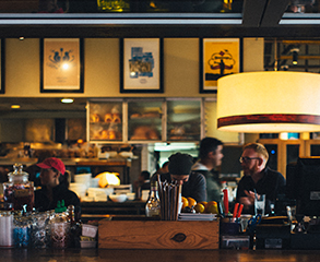 A busy restaurant interior with a view to the kitchen. There are paintings hanging near the ceiling, a bar.