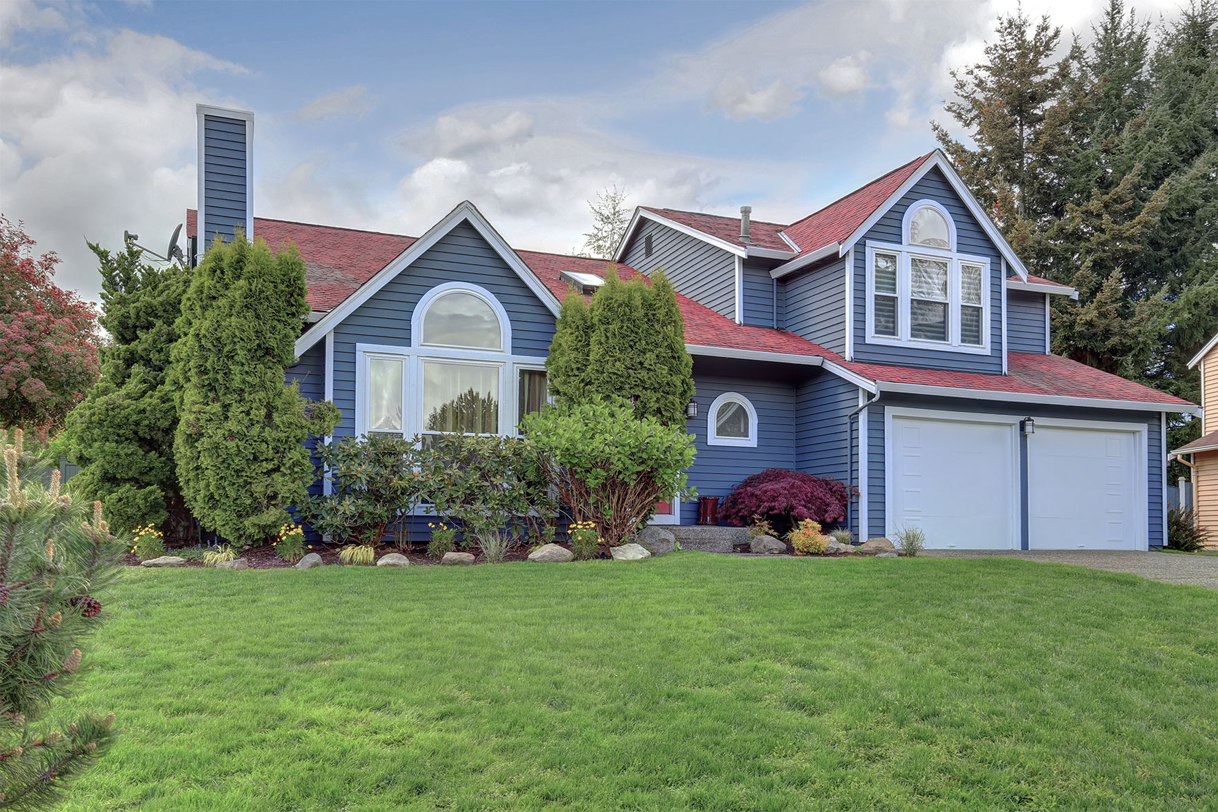 A house with blue exterior paint, bright red roof, large white windows and tidy landscaping.