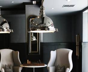 Luxury interior with two grey armchairs, a table with a chess board, large metal lamps and and a modern mirror hung on a half grey and half black painted wall.