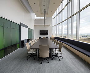 A clean and bright conference room in an office with a flat screen TV, long conference table, 16 chairs around it and big windows that allow sunlight in.
