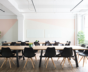 A large office space with white and pink colored walls, large windows, two long wood tables and a lot of black chairs around them.