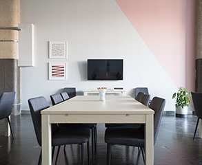 A modern office space with white and pink painted walls, large wood table with six chairs in the center, a flatscreen TV and two small paintings on the wall, and a plant in the corner of the room.