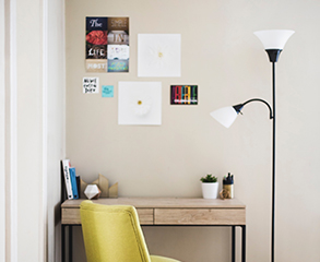 A beige home office in a condo. The walls are painted a clean white, there are several pictures hanging above the small wood desk, there are some books and two plants on it, a yellow chair is next to the desk, and there is a white lamp right to the desk.