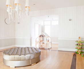 White interior with light wood floors, a large ottoman in the center of the room and kid’s toys.