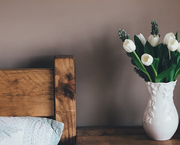 A fragment of a wood bed and a bedside table with a vase of flowers on it, in front of a brown wall.