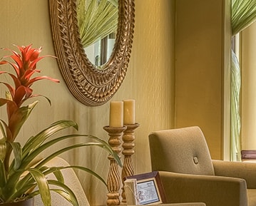 A fragment of a yellow painted living room with wooden candle holders, light brown armchairs, a framed mirror and a plant.