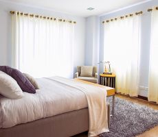 An airy bedroom with light blue walls, white curtains, fluffy blue carpet, a black and white armchair  in the corner  with a yellow and black end table next to it and in the center of the room there is a large white double bed with four cushions on it.