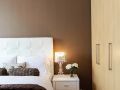 White bed, nightstand with a lamp and flowers on it, and a closet in front of a brown wall painted by Pro Painters.