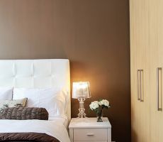 White bed, nightstand with a lamp and flowers on it, and a closet in front of a brown wall painted by Pro Painters.