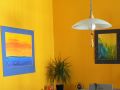 Corner of a mustard colored room with two paintings hanging on the adjoining walls, a ponytail palm on the table and a large hanging light.