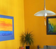 Corner of a mustard colored room with two paintings hanging on the adjoining walls, a ponytail palm on the table and a large hanging light.